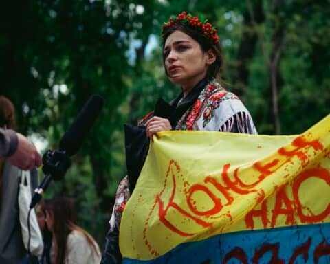 A woman holds a bloodstained flag of Ukraine in her hands, dressed in traditional Ukrainian clothing, protesting the war. Someone is holding a microphone close to her.